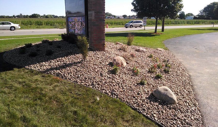Landscaped area with rocks, small plants, and a signpost next to a road with passing cars.