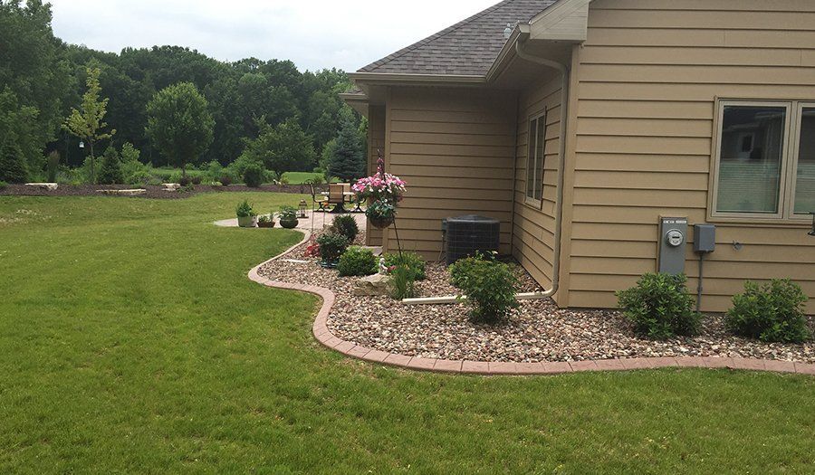 A house with tan siding and a gravel landscape bed edged with a red brick border, green grass.
