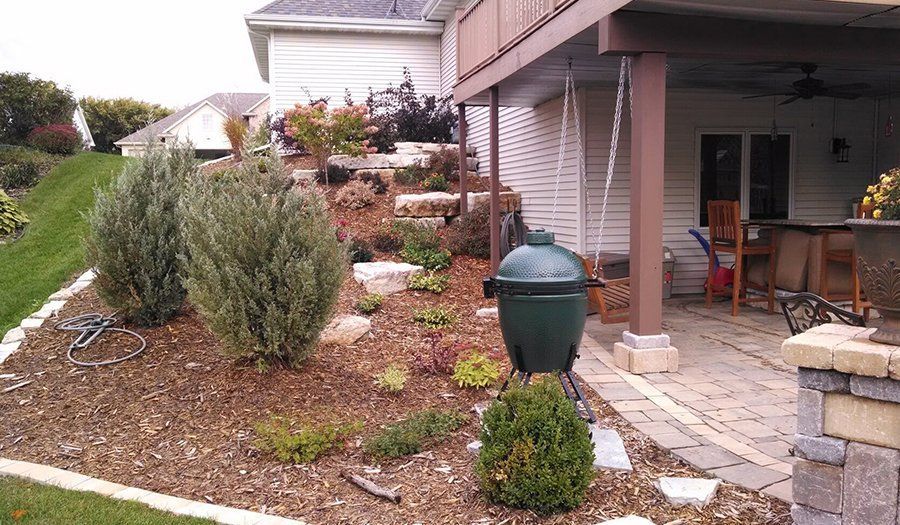Landscaped yard with stone patio, green Big Green Egg grill, and raised garden beds.
