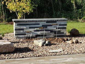 Water fountain with layered stone facade in a gravel bed, with grass and trees in the background.