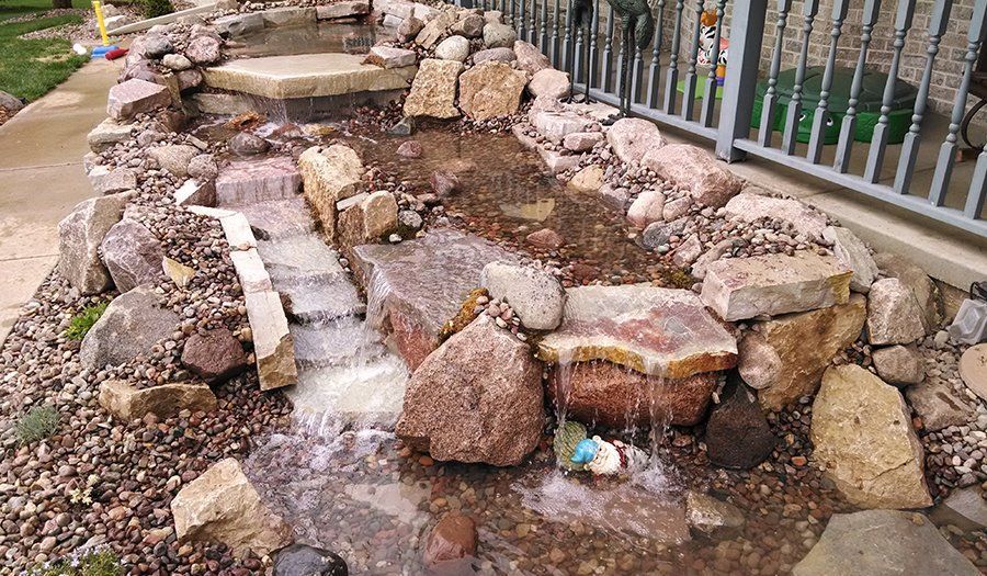 A small, rocky waterfall feature in a garden, with water cascading down rocks and a nearby fence.
