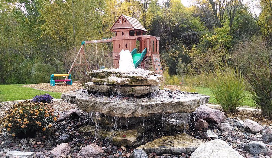 Fountain with tiered stone structure; in background, a wooden playhouse and swing set on a grassy lawn.