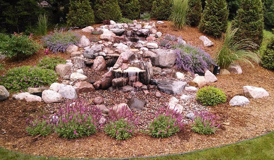 Water cascading down rocks in a landscaped garden bed with plants, mulch, and surrounding greenery.