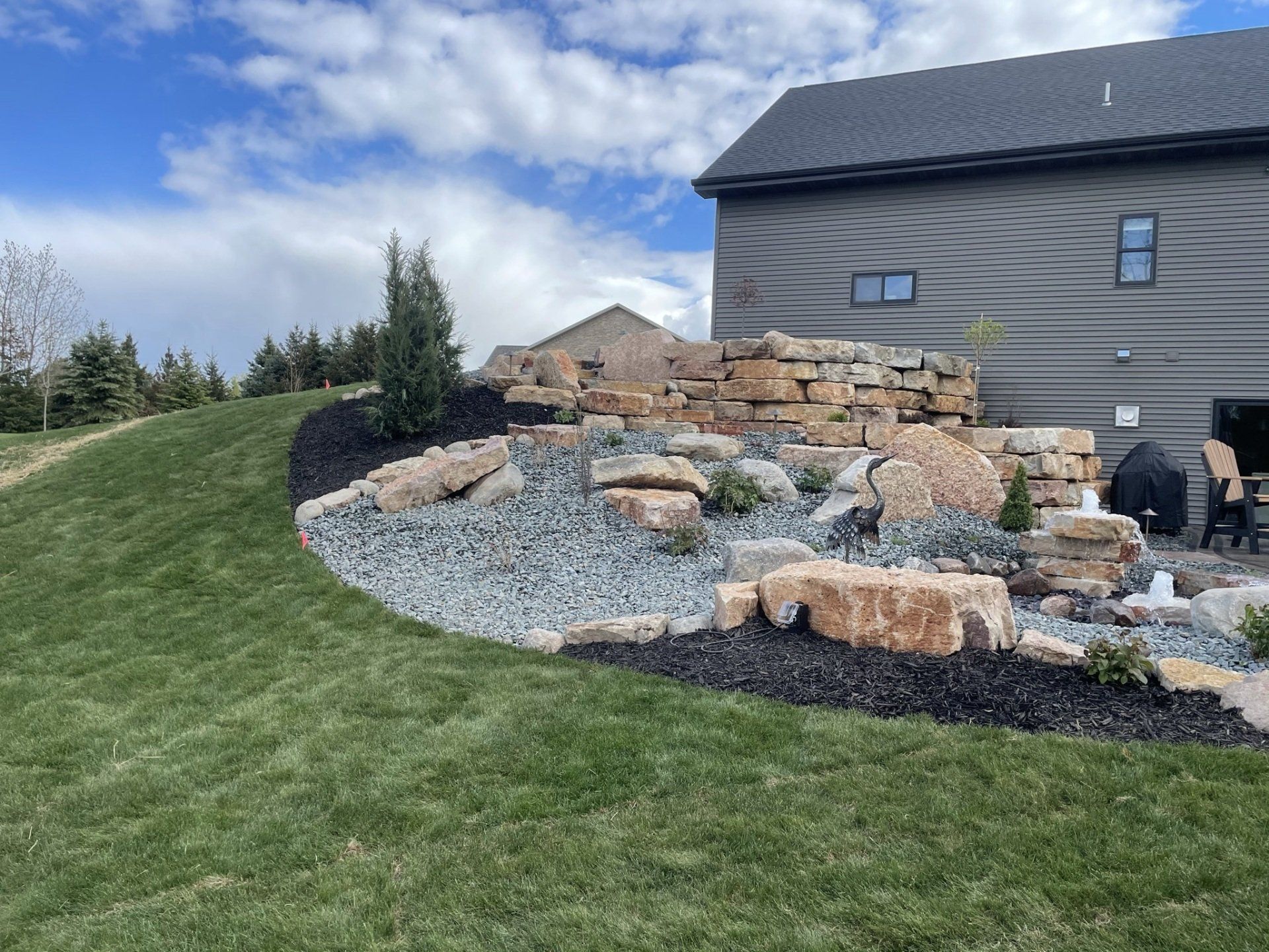 Backyard landscape with rock features, mulch, and green lawn against a gray house and blue sky.