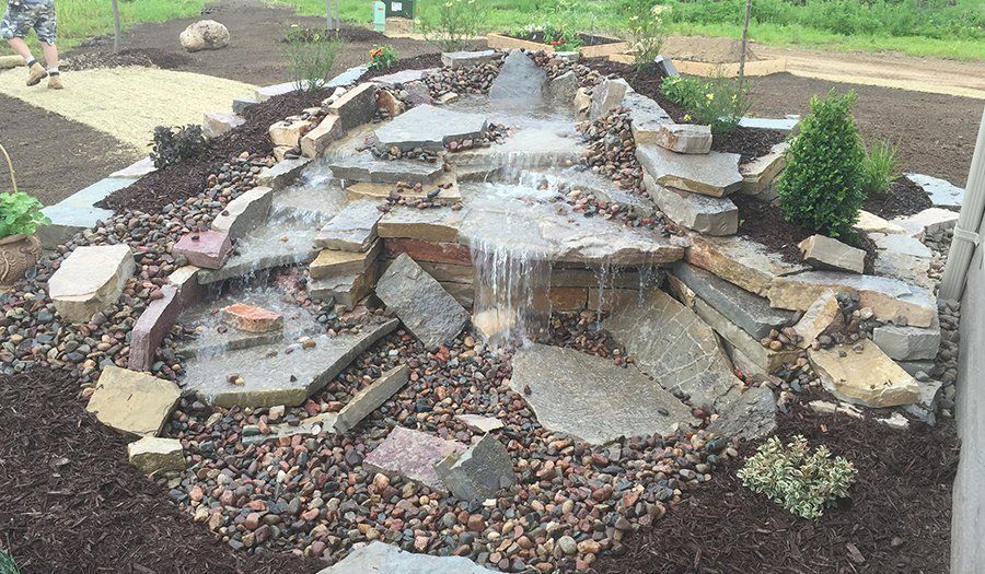 Water feature with stacked stone waterfall cascading into a gravel-lined basin, surrounded by mulch and greenery.