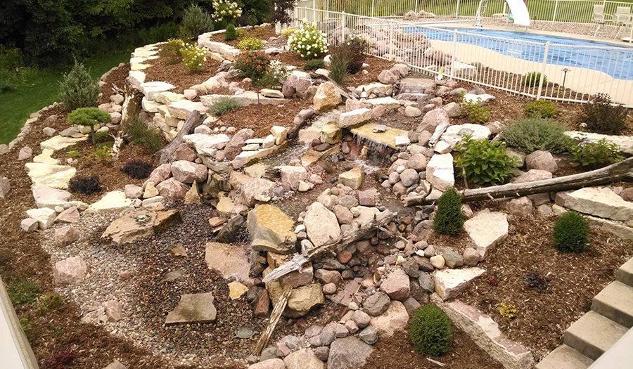 Water feature with rocks, plants, and small waterfall near a pool.