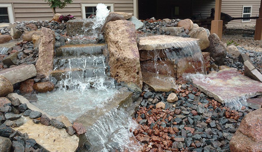 Water cascading down a multi-tiered rock waterfall feature, flowing into a bed of dark and red stones.