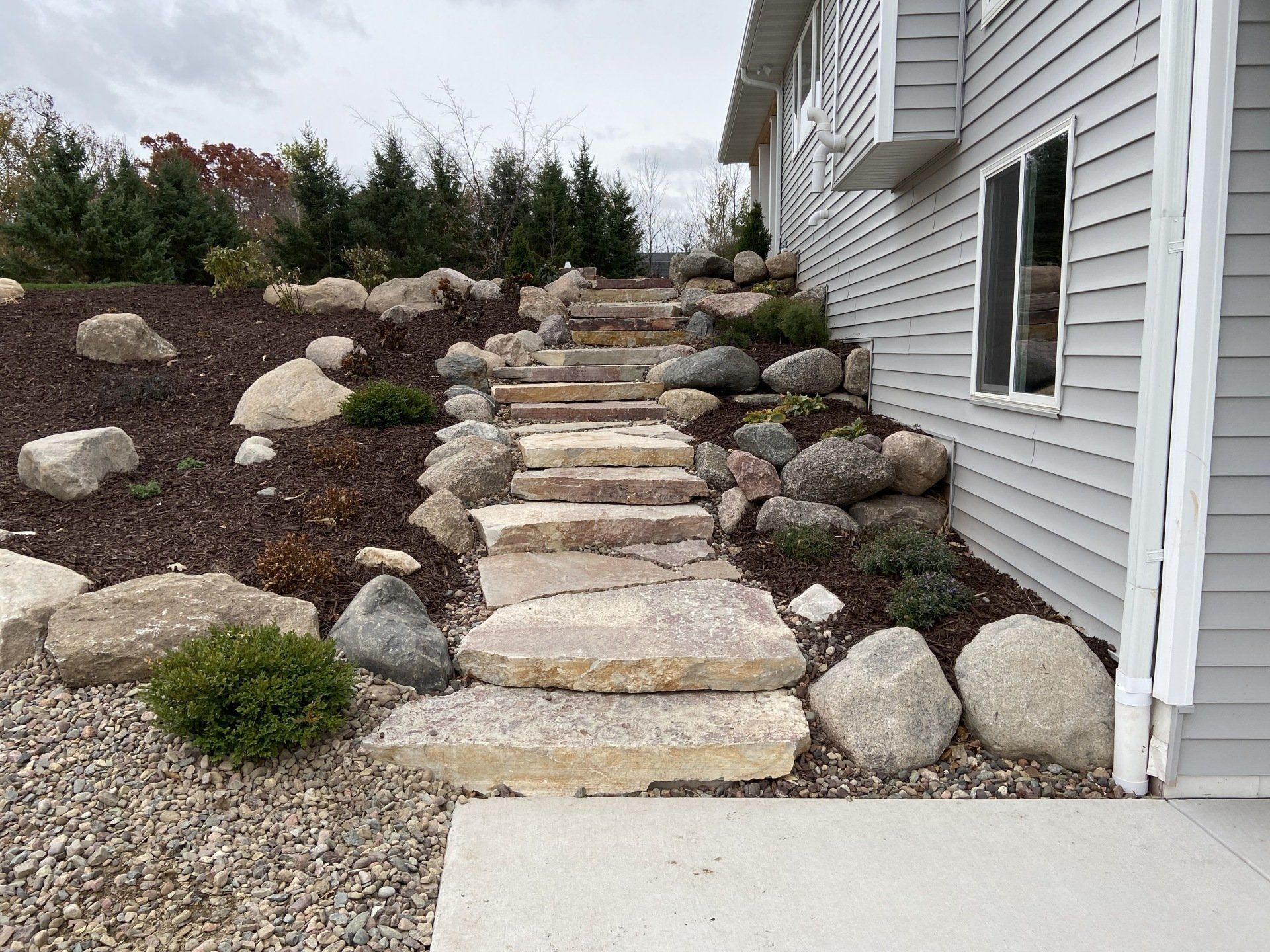 Stone steps leading up a hillside, beside a house with gray siding.