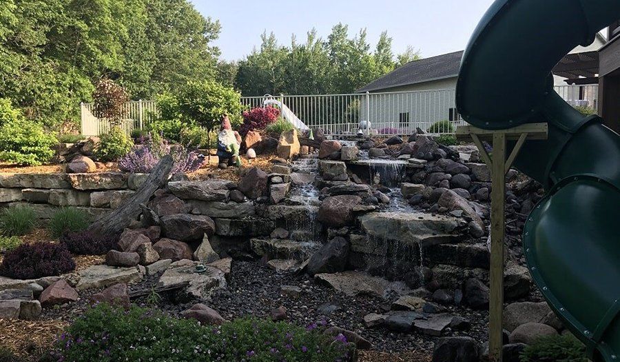 Backyard with a rock waterfall feature, greenery, and a green slide.