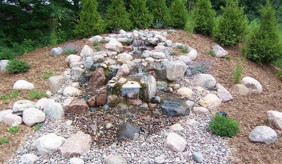 Water cascading down a rocky waterfall feature in a garden, surrounded by small trees and wood chips.
