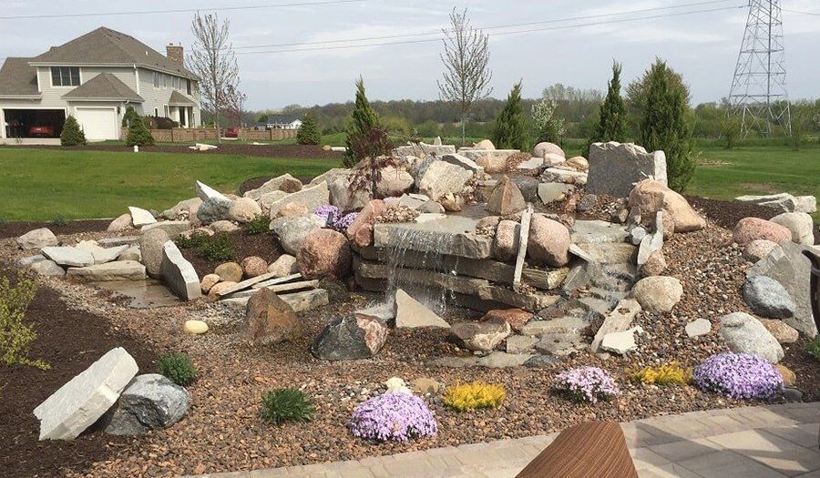 A backyard waterfall feature with various sized rocks, mulch, and colorful flowers, in front of a house.