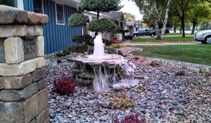 Water fountain in a landscaped yard with a rock border, next to a blue house and mailbox pillar.