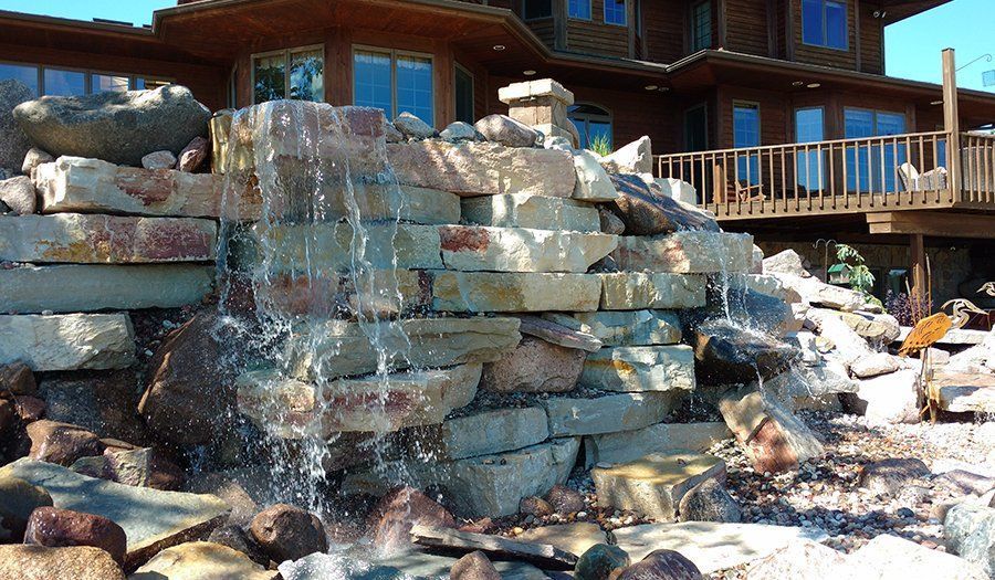 Water cascades over a stone waterfall in front of a multi-story wood home.