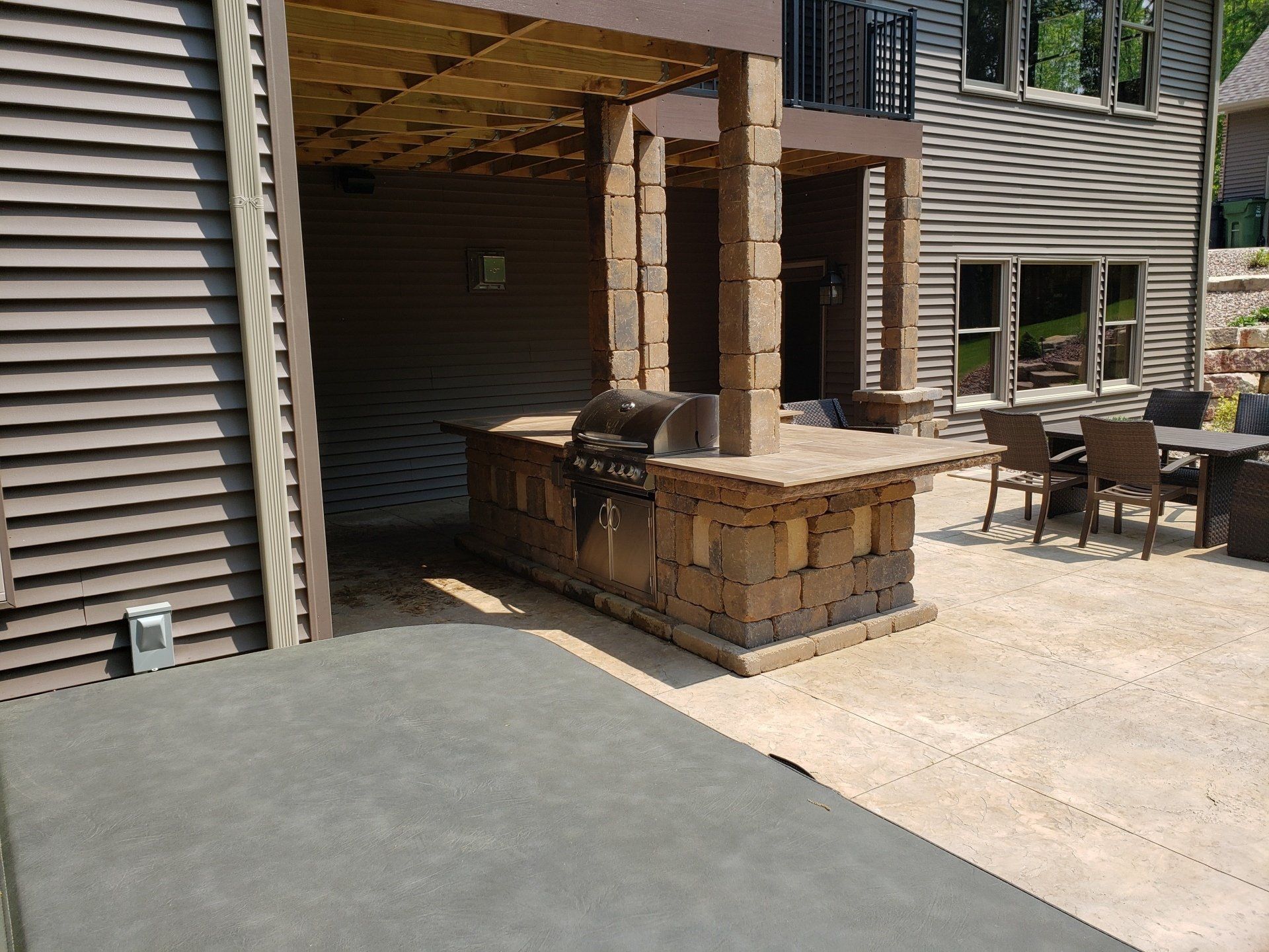 Outdoor kitchen with grill, countertop, and seating area. Brown and tan stone, concrete patio, and home exterior.