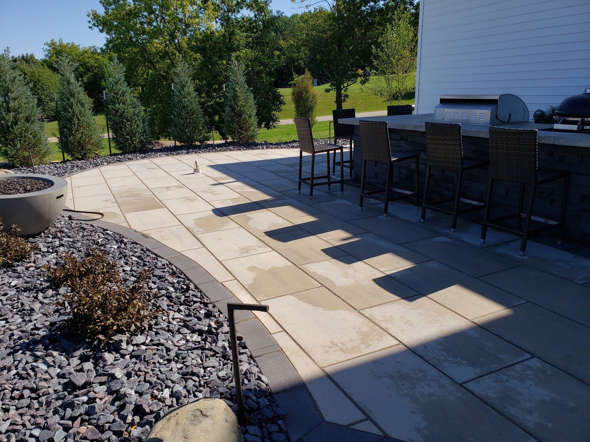 Patio with stone pavers, outdoor bar with stools, a fire pit, and landscaping under a sunny sky.
