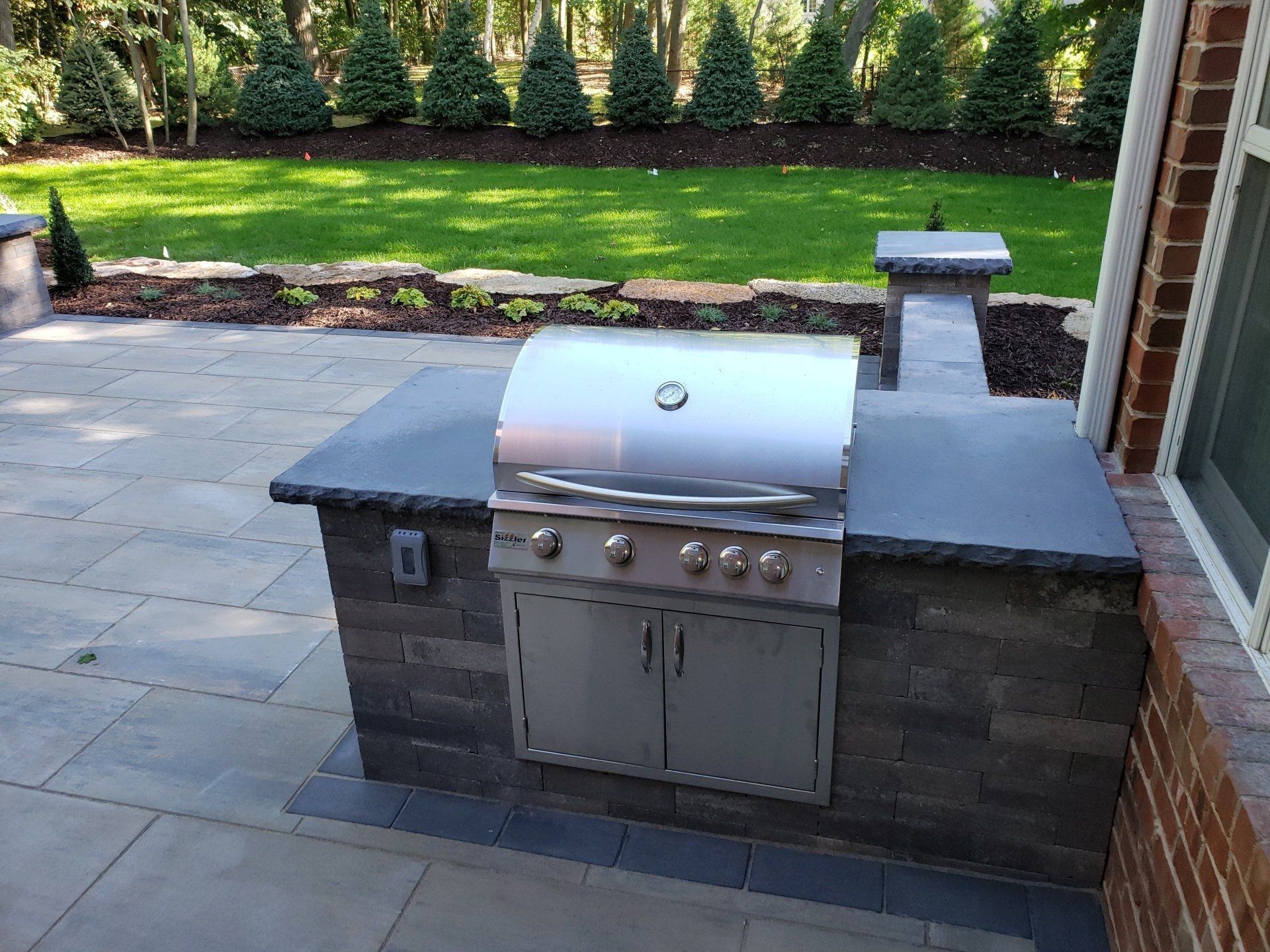 Outdoor kitchen with grill built into a stone island on a patio, with a grassy lawn in the background.