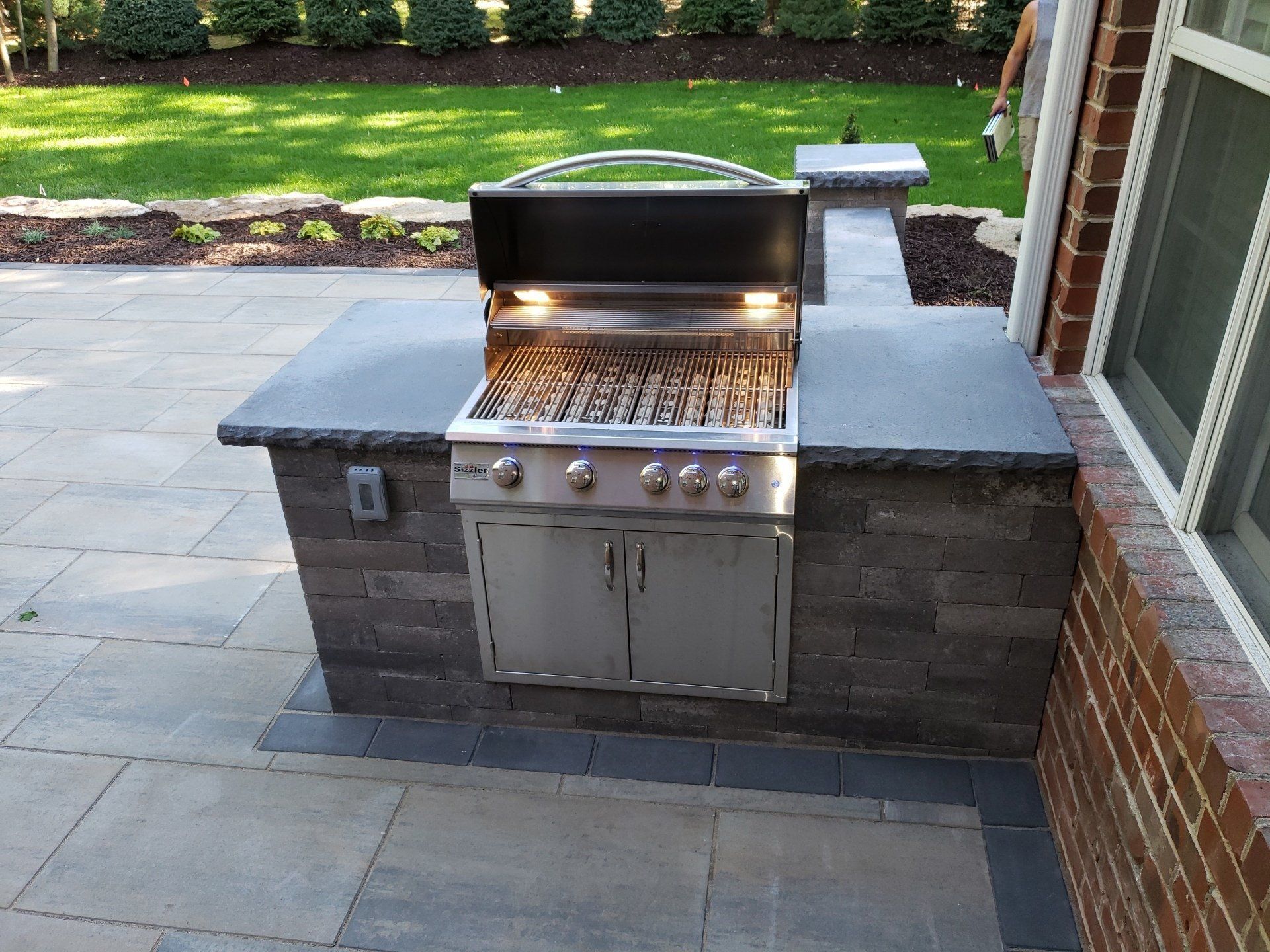 Outdoor built-in barbecue grill with stainless steel doors and a gray stone countertop on a patio.