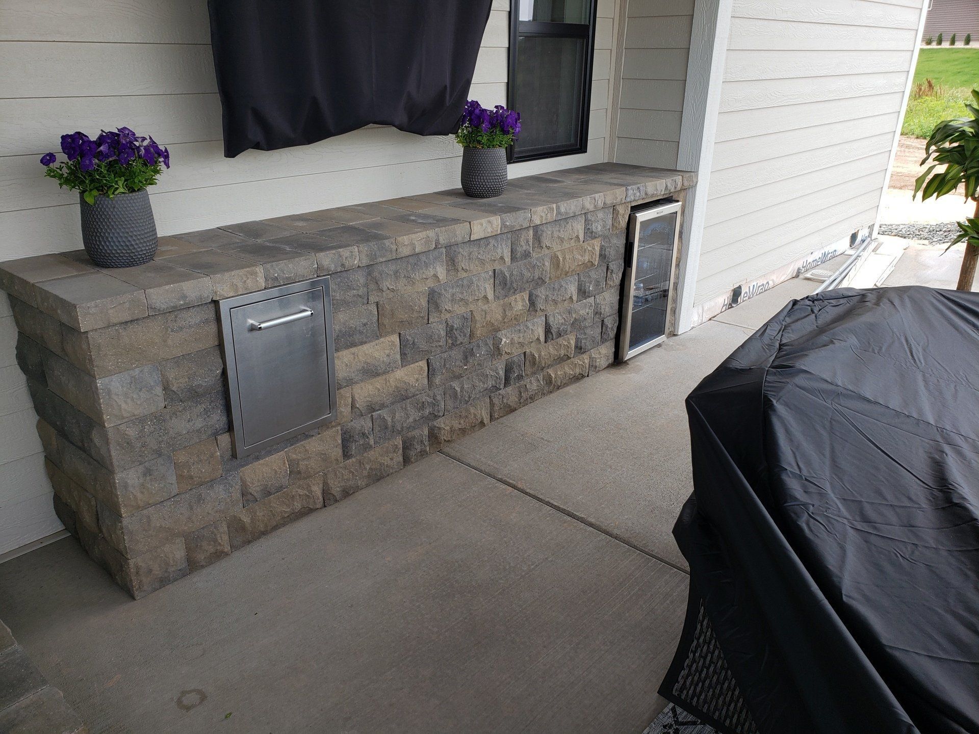 Stone-faced outdoor counter with a mailbox, a door, and two potted plants with purple flowers. A covered grill is visible.