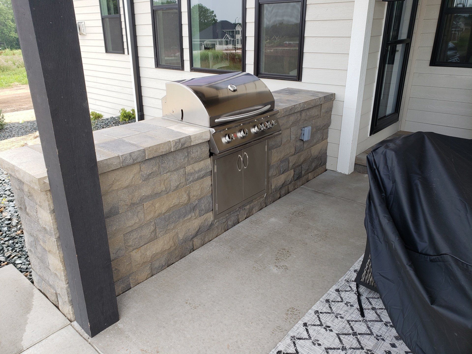 Built-in outdoor grill with stone surround, on a concrete patio, next to a covered porch.