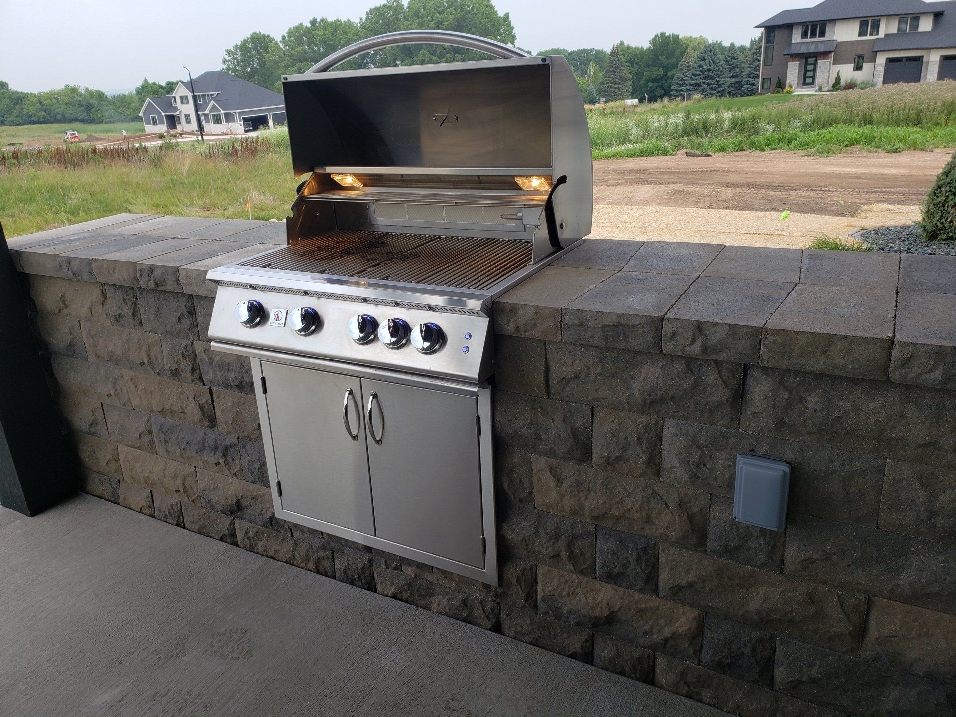 Outdoor stainless steel grill built into a stone wall. Flames visible.