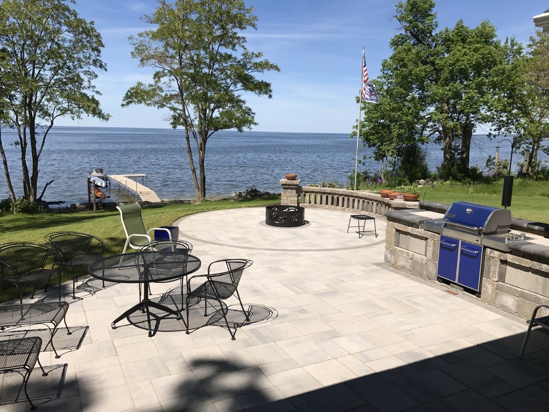 Patio overlooking a lake with outdoor seating, a grill, and a fire pit. Trees and a dock in the background.