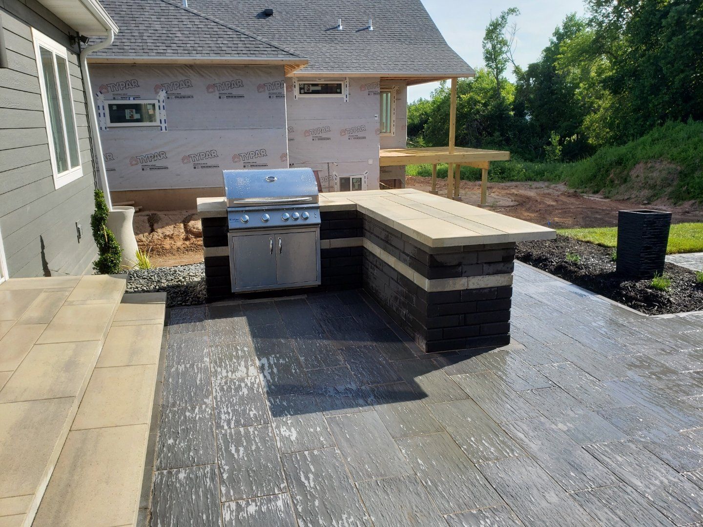 Outdoor kitchen with a grill and countertop, built on a gray stone patio near a house under construction.