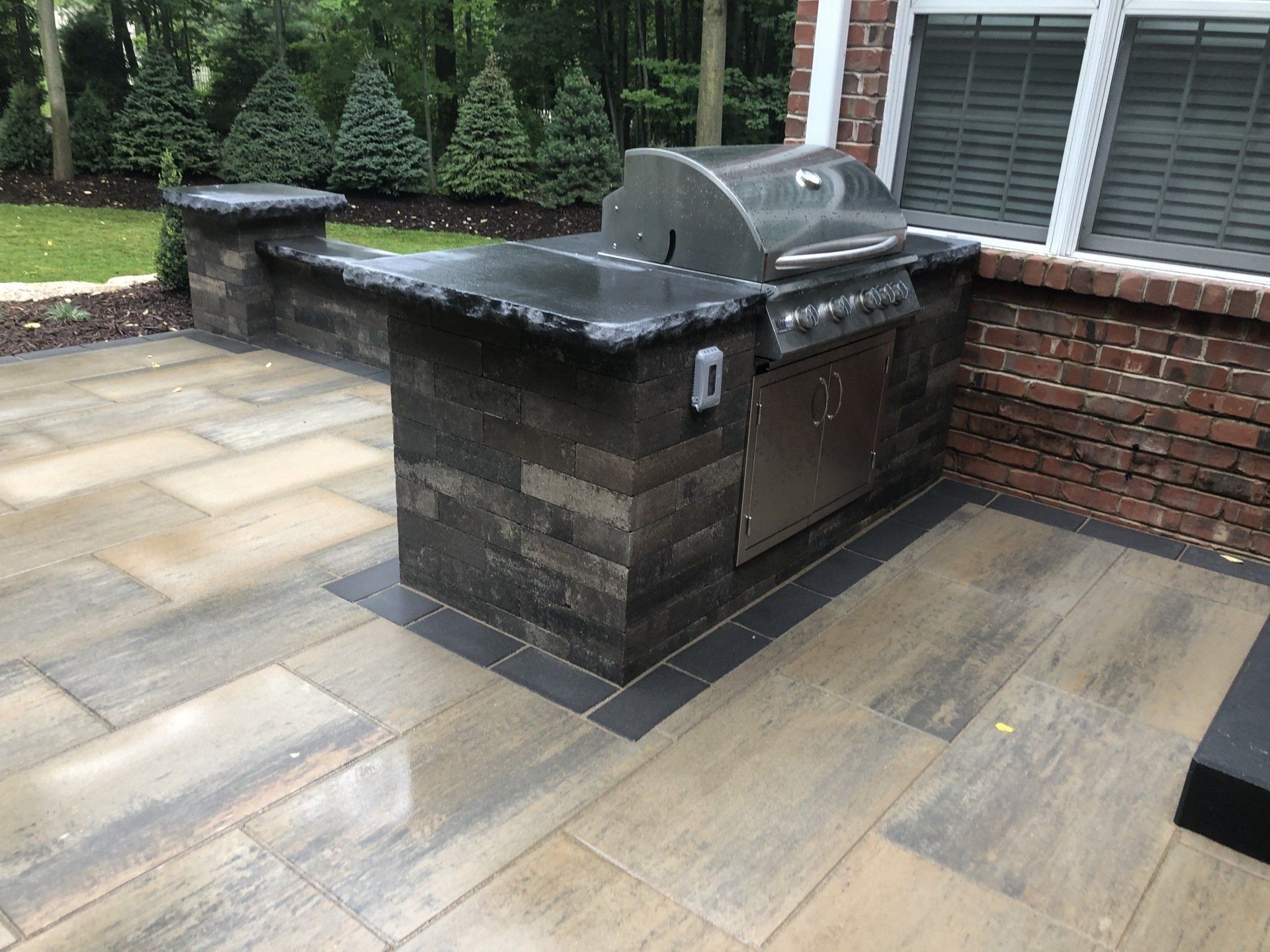 Outdoor kitchen with a grill, countertop, and brick accents on a stone patio.