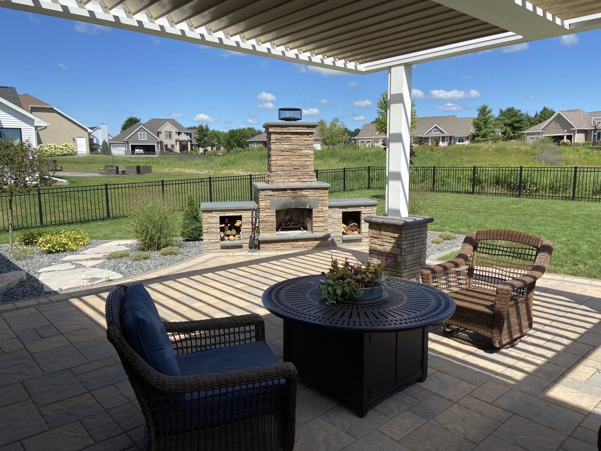 Patio with a fireplace, chairs, and table under a pergola, overlooking a grassy yard with houses.