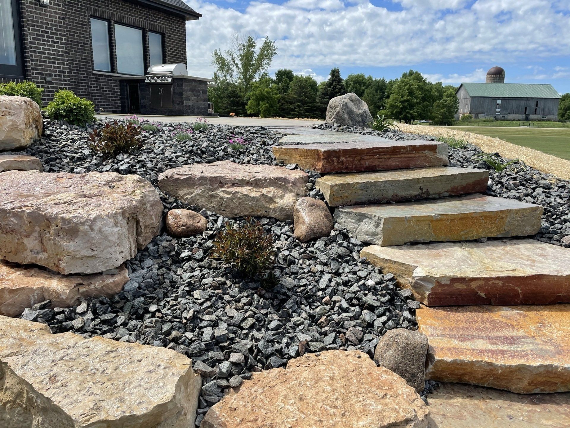 Stone steps and large rocks ascend a landscaped area with dark gravel, shrubs, and a house in the background.