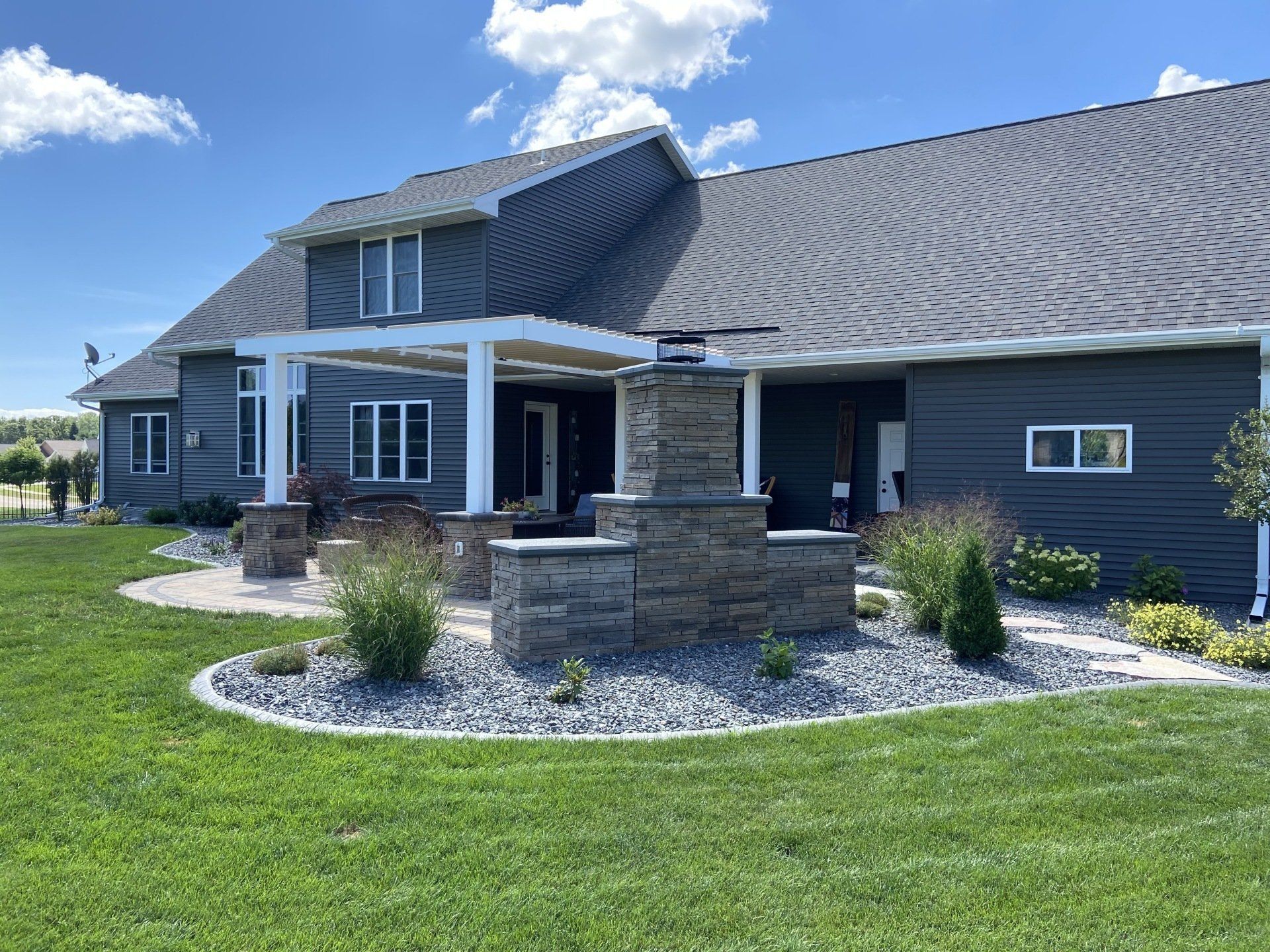 Exterior of a modern home with dark blue siding, covered patio, stone fireplace, and well-maintained landscaping.