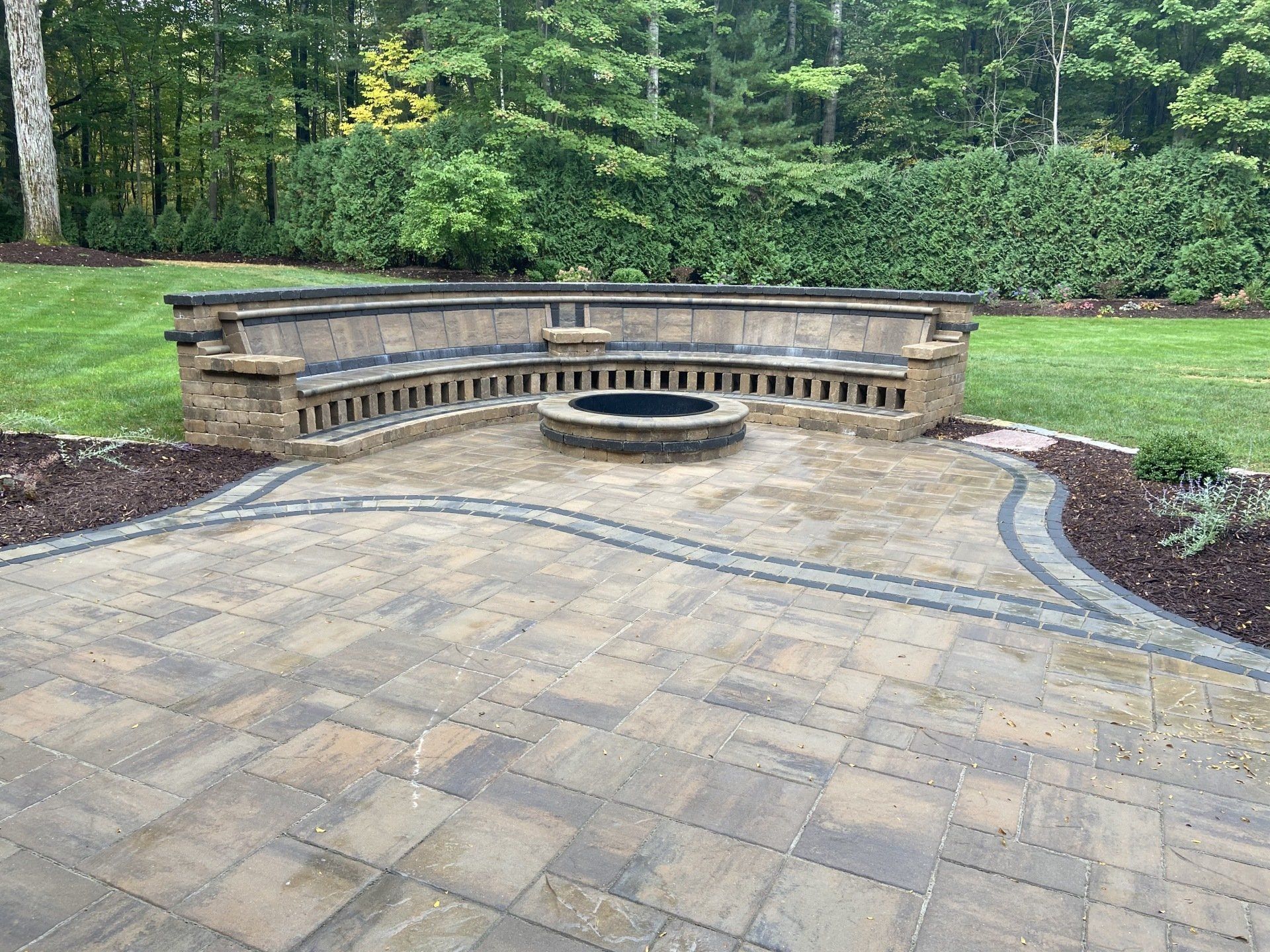 Outdoor patio with built-in stone seating around a fire pit, with surrounding greenery and lawn.