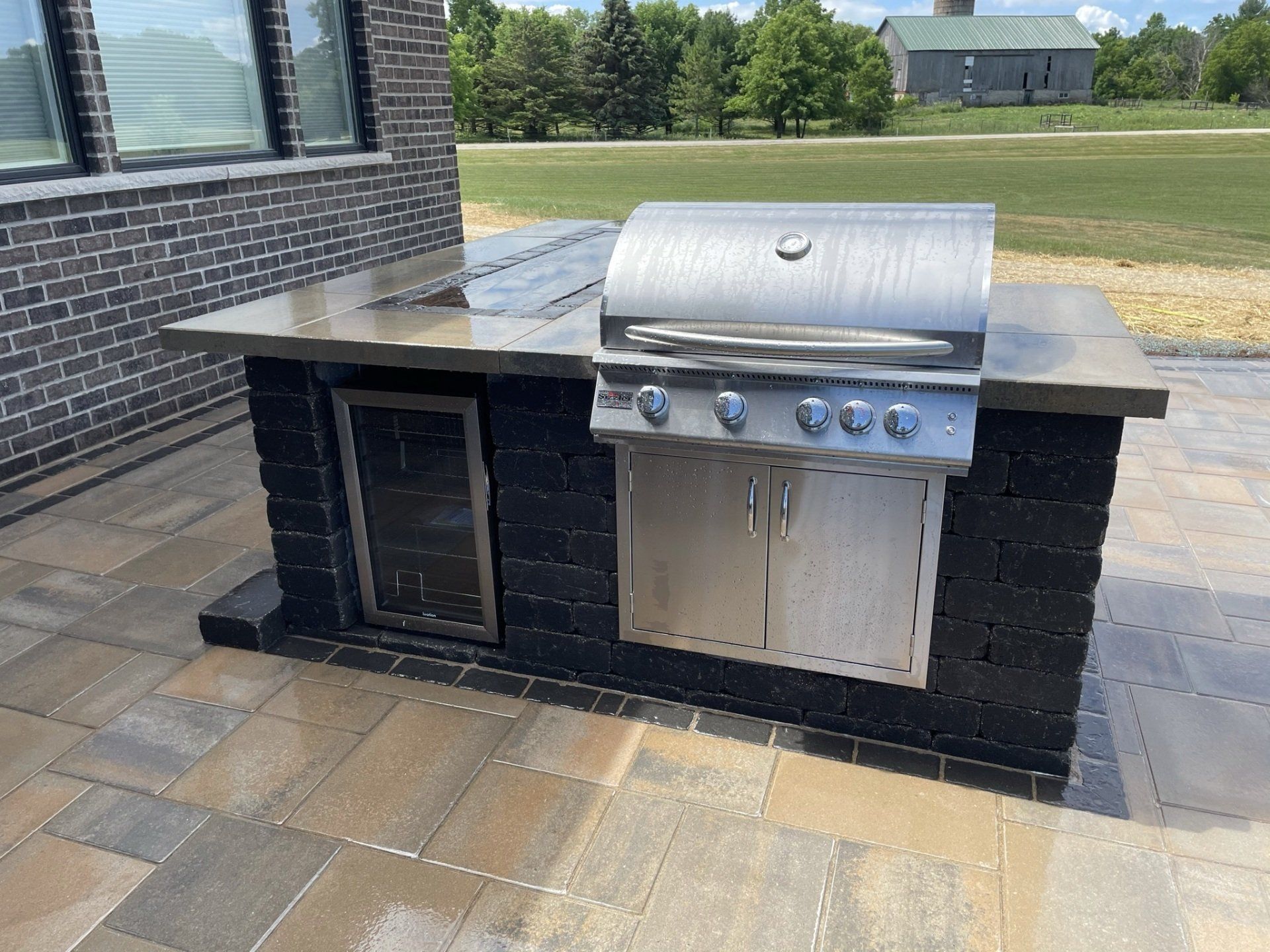 Outdoor grilling station with stainless steel grill, beverage cooler, and dark brick base on patio.
