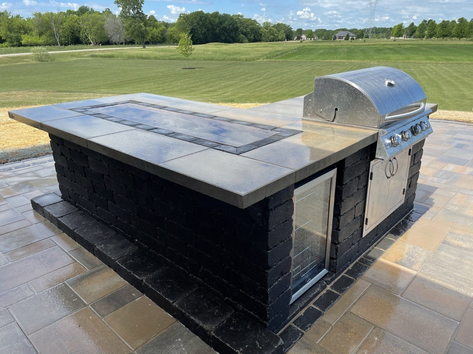 Outdoor kitchen island with grill and fire pit, on a paved patio with green grass in the background.