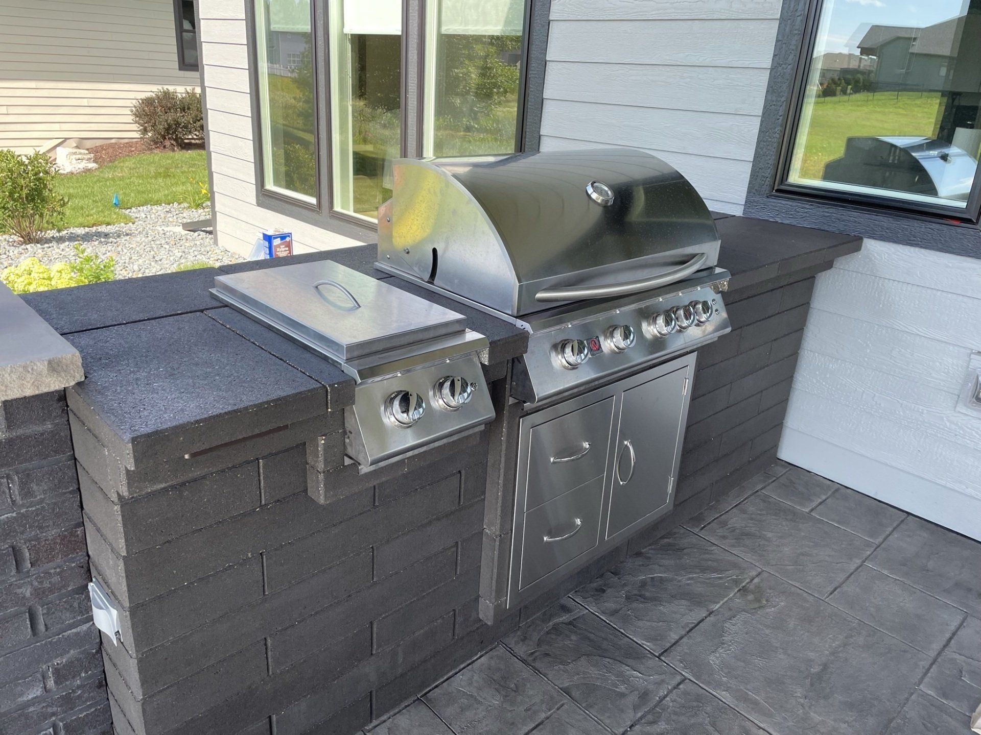 Stainless steel outdoor grill and side burner built into a dark gray brick counter on a patio.