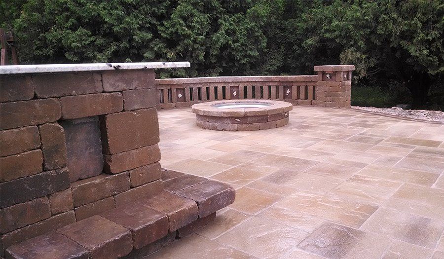 Stone patio with built-in fireplace, fire pit, and decorative railing. Green trees in the background.