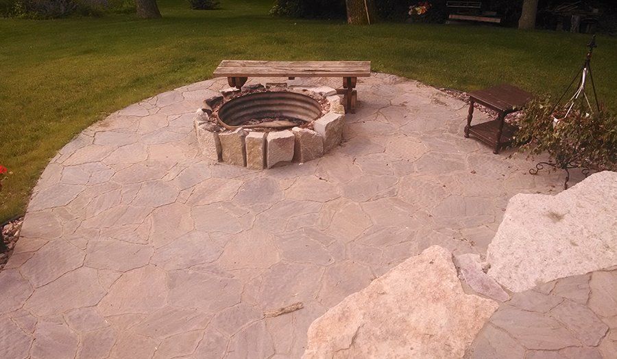 Stone fire pit with bench and additional seating on a paved patio, surrounded by green grass.