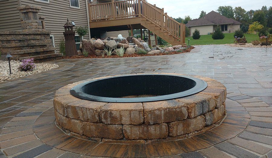 Brick fire pit on a patterned brick patio, with a house and deck in the background.