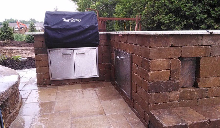 Outdoor kitchen with brick facade and stainless steel grill, cabinets, and stone patio.