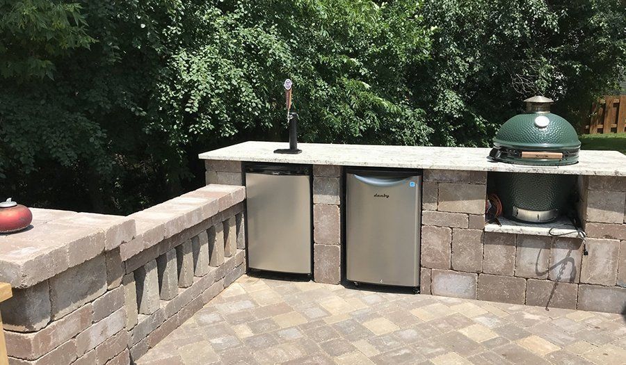 Outdoor kitchen with a grill, two refrigerators, and a tap, set in a brick patio with lush greenery.