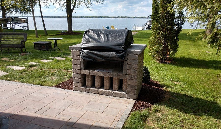 Brick grill on a patio, covered with a black tarp, near a lake.