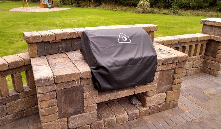 Built-in brick barbecue with a black cover on a patio, with a grassy lawn in the background.
