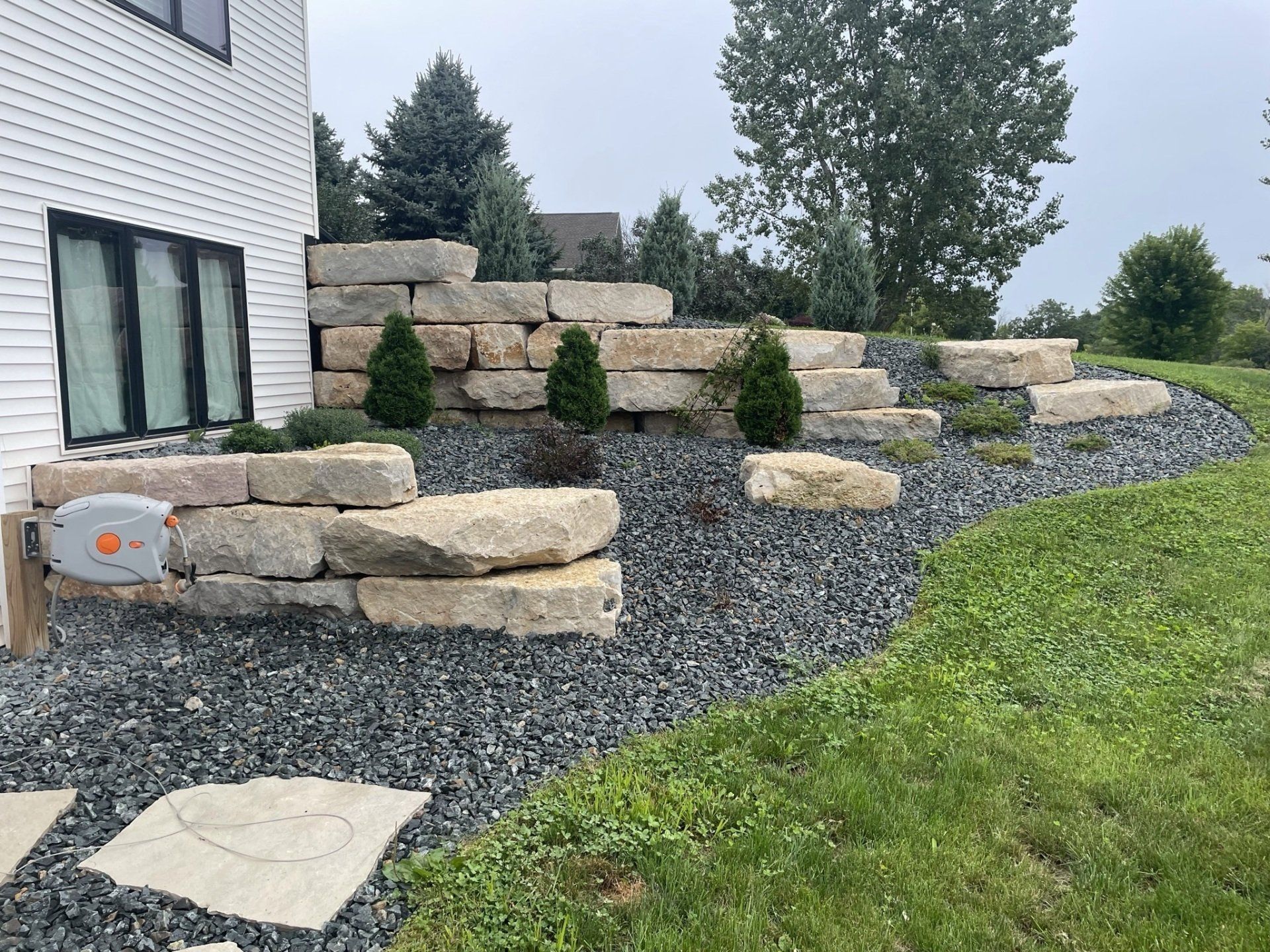 Stone retaining wall with steps, black gravel, small trees, and green grass.