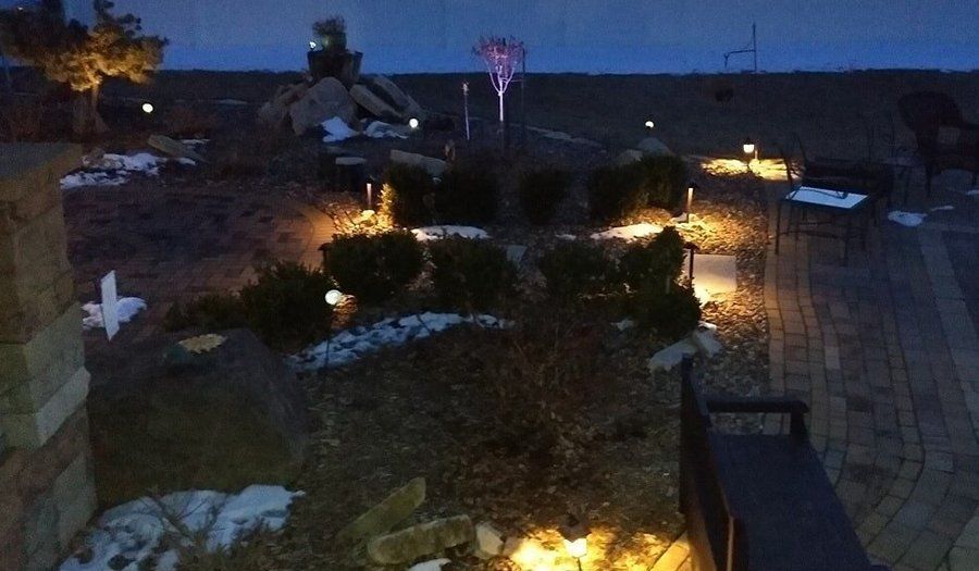 Lit outdoor landscaping at dusk. Small lights illuminate bushes, rocks, and a cross-shaped decoration.