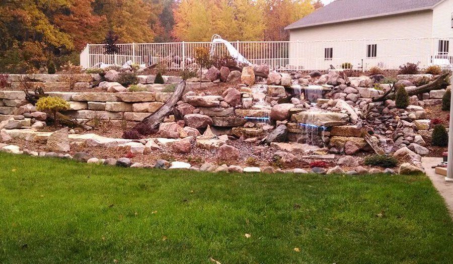 A tiered rock garden with waterfall, lush green grass, and a white house in the background.