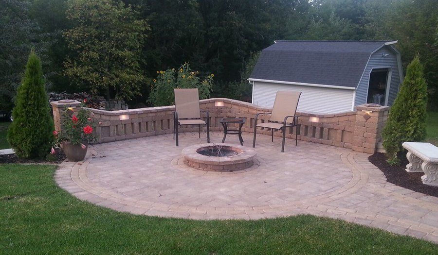 A brick patio with fire pit, two chairs, and a small shed in the background. Green grass surrounds the patio.