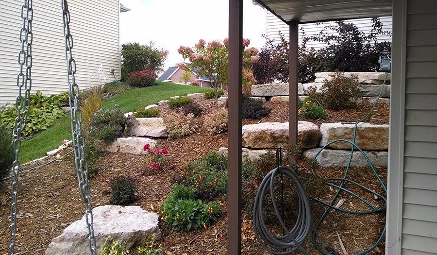 A tiered garden with stone steps and various plants, seen next to a house and under a covered area.
