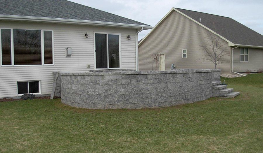 Backyard patio with retaining wall, steps, and two-story houses. Gray blocks, green grass, and cloudy sky.
