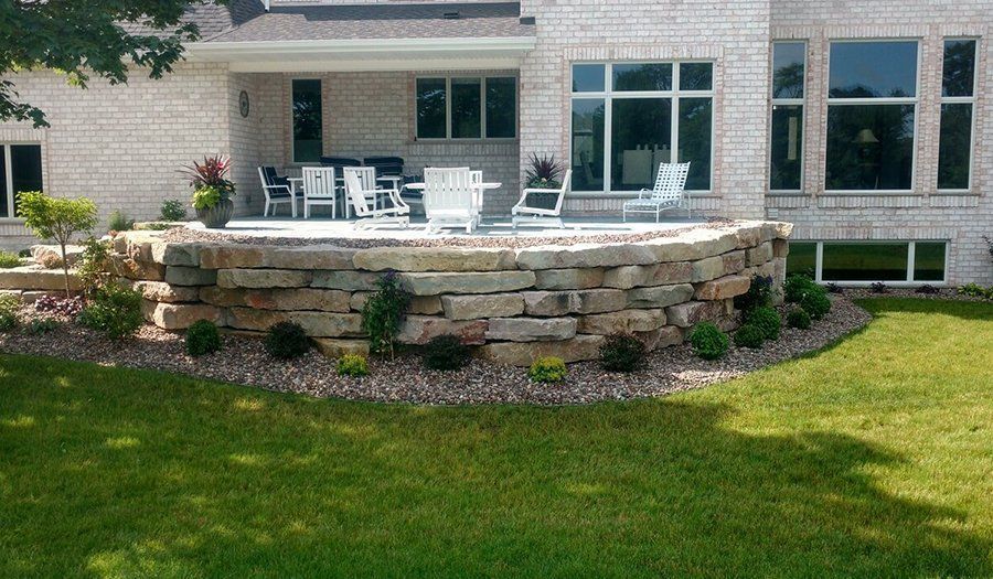 Raised stone patio with white chairs and landscaping near a brick house.