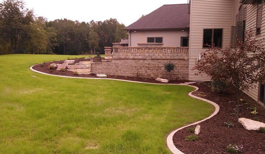 Green lawn curving around a stone patio and house with a brown roof and siding.