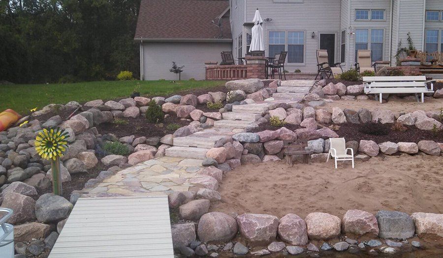 Stone steps and landscaping lead from a lake dock to a house with outdoor seating.